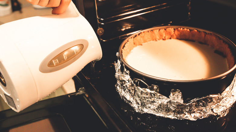 Κάποιος ρίχνει νερό από ένα βραστήρα σε ένα bain-marie που κρατά ένα cheesecake στο φούρνο