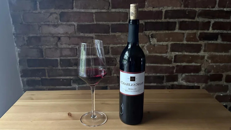 Ένα ποτήρι Charles Shaw Merlot κάθεται σε ένα τραπέζι δίπλα σε ένα ανοιχτό μπουκάλι.